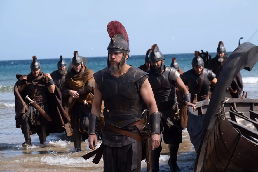 Matt Damon as Odysseus, in a leather cuirass and a helmet crowned with a tall red horsehair crest, walks up the beach ahead of his soldiers hauling their ship out of the surf.