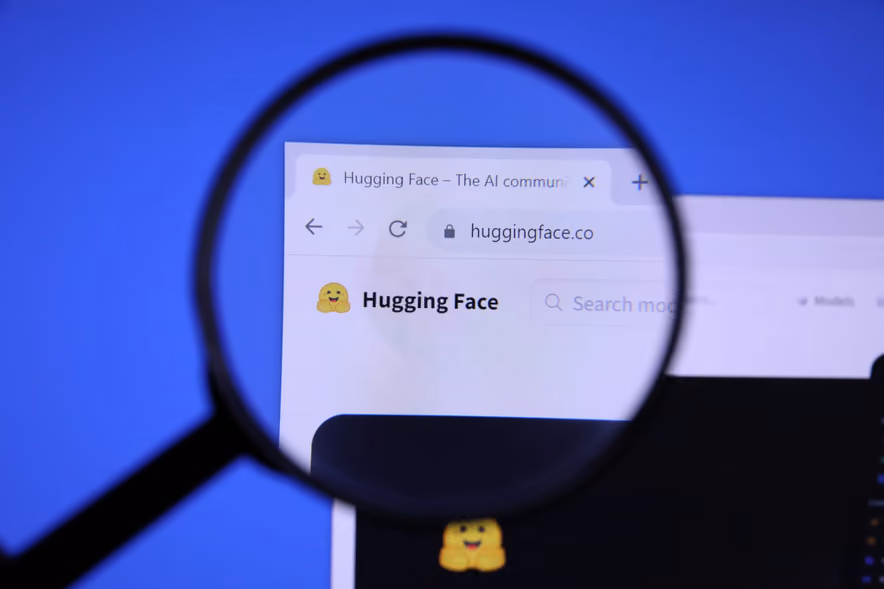A magnifying glass held in front of a screen enlarges the header of huggingface.co, the yellow open-armed smiling emoji logo next to the Hugging Face name, against a blue background.