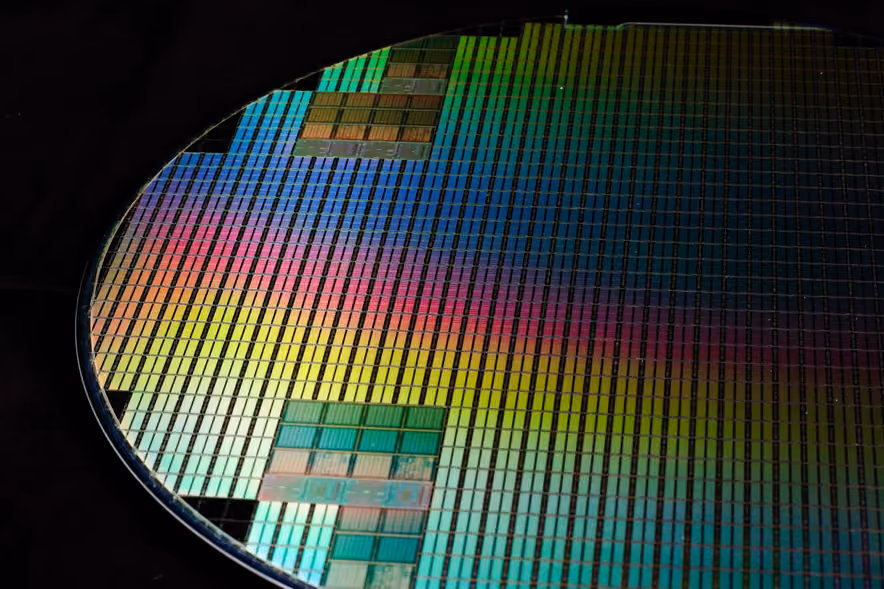 Close-up of a tilted silicon wafer covered in hundreds of rectangular chips, throwing iridescent green, pink, and blue reflections against a black background.