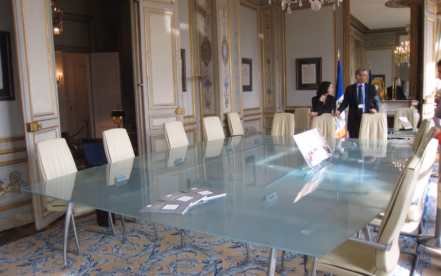 The meeting room of France's Constitutional Council, a long frosted-glass table ringed with cream leather chairs under a crystal chandelier, between white and gold paneling and a blue arabesque carpet.