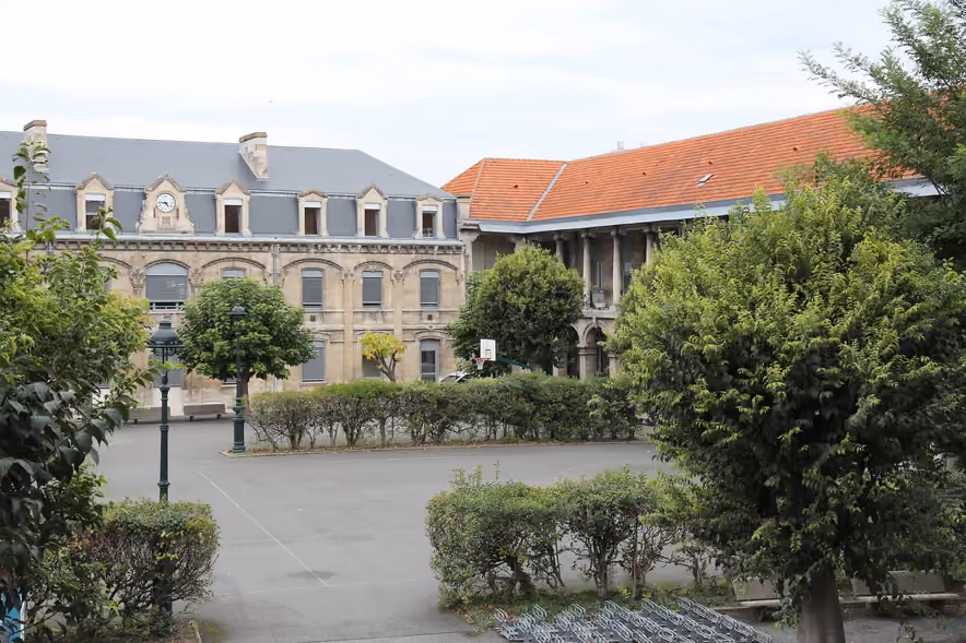 An empty high school courtyard, a wide paved yard edged with hedges and shrubs, facing a pale stone building topped by a clock and a long wing under orange tiles, with a basketball hoop and empty bike racks.