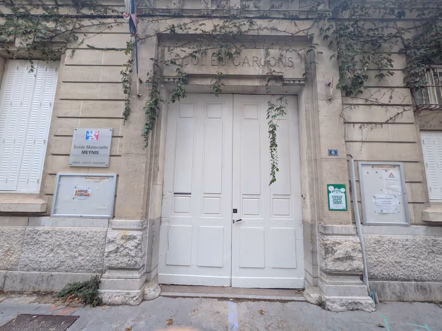 Stone facade of a former boys' school turned preschool, with a tall white double door, ivy trailing from the lintel, a plaque reading "École maternelle Meynis," and two glass-fronted notice boards filled with sheets of paper.