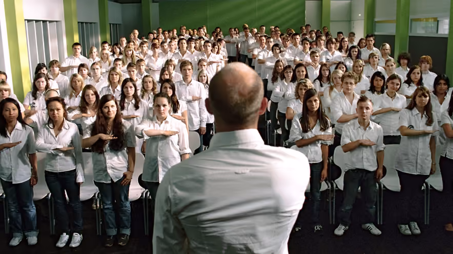 Facing a teacher seen from behind in a white shirt, an entire room of students in white shirts performs the same salute, right arm stretched across the chest.