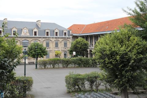Cour intérieure déserte d’un lycée, un vaste préau bitumé bordé de haies et d’arbustes, devant un bâtiment de pierre claire surmonté d’une horloge et une longue aile aux tuiles orange, avec un panier de basket et des range-vélos vides.