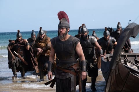 Matt Damon as Odysseus, in a leather cuirass and a helmet crowned with a tall red horsehair crest, walks up the beach ahead of his soldiers hauling their ship out of the surf.