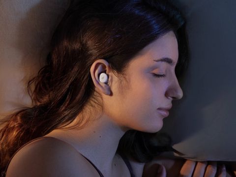 A pair of white earbuds built for sleeping, lying flat beside their oval charging case on a light background.