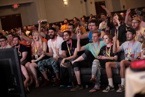 A packed convention hall of spectators sitting in the dark, facing a lit stage where a game is being played on a giant screen.
