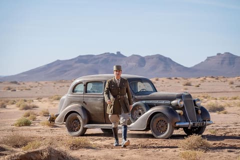 Simon Abkarian in a general's uniform and kepi, walking past a black 1940s car parked in the middle of a desert, barren mountains on the horizon.