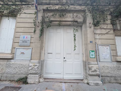 Stone facade of a former boys' school turned preschool, with a tall white double door, ivy trailing from the lintel, a plaque reading "École maternelle Meynis," and two glass-fronted notice boards filled with sheets of paper.