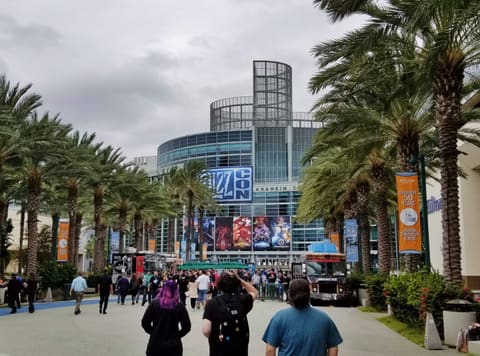 The glass facade of the Anaheim Convention Center on a BlizzCon day, the show's blue logo above posters for Warcraft, Diablo, Hearthstone, and Overwatch, palm trees on both sides, and visitors in black T-shirts walking up the promenade.