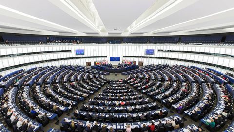 Overhead view of the European Parliament hemicycle in Strasbourg during a plenary session, members seated in concentric rings around a podium below the EU flag and the flags of the member states.