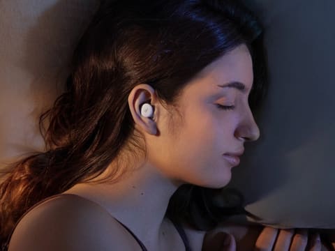 A pair of white earbuds built for sleeping, lying flat beside their oval charging case on a light background.