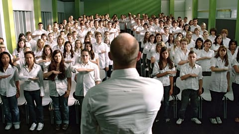 Facing a teacher seen from behind in a white shirt, an entire room of students in white shirts performs the same salute, right arm stretched across the chest.