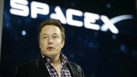 Elon Musk, wearing a headset microphone, speaks in front of a giant screen displaying the SpaceX logo against a starry sky.