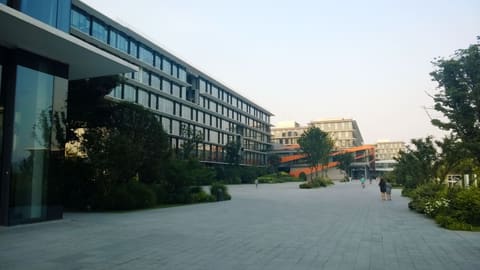 A wide paved plaza on Alibaba's Hangzhou campus, lined on the left by a long glass office building with horizontal awnings, half screened by trees. More glass buildings and an orange walkway close off the far end, where a few figures walk under a pale, hazy sky.