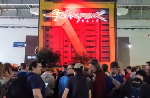 A dense crowd of convention visitors, nearly all seen from behind, waits in front of a huge booth topped by the red and white neon sign of Cyberpunk 2077, mounted on a fake apartment facade.
